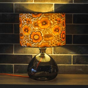 May include: A table lamp with a floral lampshade. The lampshade has a pattern of orange and brown flowers on a cream background. The base is a dark glass bottle. An orange cord extends from the base.
