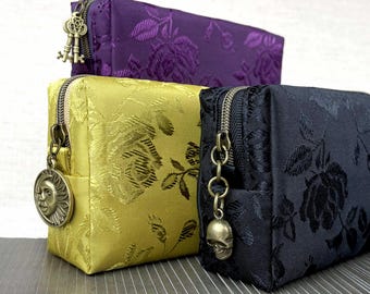 May include: Three rectangular zippered pouches in purple, gold, and black, each featuring a tonal rose pattern. The purple pouch has a key charm, the gold pouch has a sun face charm, and the black pouch has a skull charm on the zipper pull.