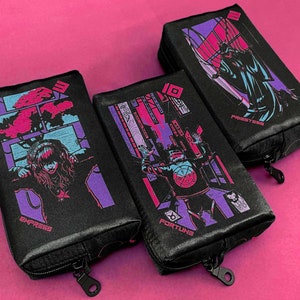 May include: Three black zippered pouches with colorful illustrations. One pouch features a woman with the word "EMPRESS", another has a figure with "FORTUNE", and the third says "PRIESTESS". The pouches have a black zipper and are on a pink background.