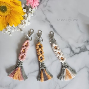 May include: Three handmade macrame keychains with a flower design. The keychains are in pink, yellow, and white with a gray tassel. The keychains are on a white marble surface. Studio Lilly Rose is written in the top right corner of the image.