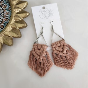 May include: A pair of blush pink tassel earrings with silver triangle frames. The earrings are handmade and feature a delicate macrame design.