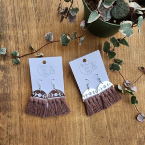 May include: Two pairs of earrings with a celestial theme. Each earring features a silver-colored, half-circle design with moon phases and a fringe of brown macrame. The earrings are displayed on white cards with the text "Studio Lily Rose".