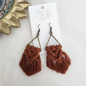 May include: A pair of handmade macrame earrings. The earrings feature a triangular brass frame with woven rust-colored cotton. The earrings have a fringe of rust-colored cotton. A white card with the text "Studio Lilly Rose" is behind the earrings.