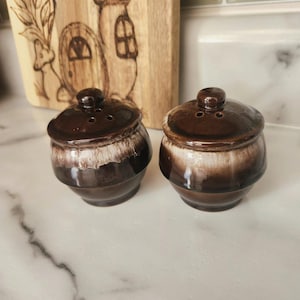 May include: Two brown and white ceramic salt and pepper shakers with a glazed finish. The shakers have a round body and a small lid with holes for dispensing salt and pepper.