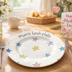 May include: A white ceramic plate with the text "Mum's lunch plate" and colorful star designs. A Mother's Day card, heart-shaped cookies, flowers, and a cup of tea are also on the table.