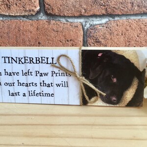 May include: Wooden memorial plaque with the text "TINKERBELL You have left Paw Prints on our hearts that will last a lifetime". A photo of a black dog is on the right side of the plaque.