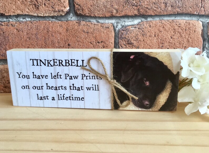 May include: Wooden memorial plaque with the text "TINKERBELL You have left Paw Prints on our hearts that will last a lifetime". A photo of a black dog is on the right side of the plaque.