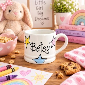 Op de afbeelding: Een witte keramische mok met de naam Betsy in zwarte schrijfletters, versierd met een gele ster links, een roze ster rechts en een lichtblauwe ster aan de onderkant. De mok heeft een afgerond handvat en een gladde, glanzende afwerking.