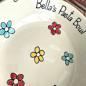 May include: A cream-coloured ceramic pasta bowl featuring hand-painted flowers in red, yellow, and light blue with black outlines. The text "Bella's Pasta Bowl" is written in black script along the upper rim of the bowl's interior surface.