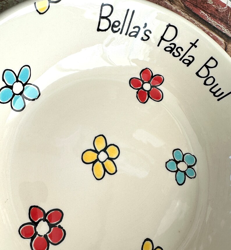 May include: A cream-coloured ceramic pasta bowl featuring hand-painted flowers in red, yellow, and light blue with black outlines. The text "Bella's Pasta Bowl" is written in black script along the upper rim of the bowl's interior surface.