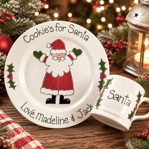 May include: A white ceramic plate and matching mug set featuring a hand-painted Santa Claus illustration. The plate reads Cookie's for Santa and Love, Madeline & Jack. The mug features the word Santa and green holly leaf accents.