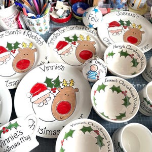 May include: A collection of white ceramic dinnerware including plates, bowls, and mugs featuring hand-painted illustrations of Santa Claus, reindeer, and holly leaves. Several pieces are personalized with names and holiday-themed text.