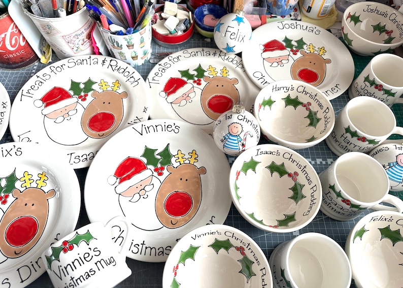 May include: A collection of white ceramic dinnerware including plates, bowls, and mugs featuring hand-painted illustrations of Santa Claus, reindeer, and holly leaves. Several pieces are personalized with names and holiday-themed text.