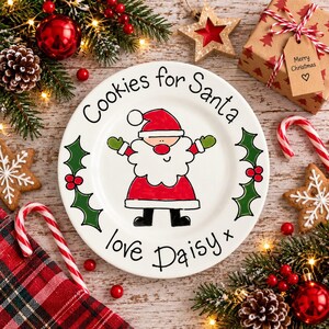 May include: A white ceramic plate featuring a hand-drawn illustration of Santa Claus in the center, surrounded by green holly leaves and red berries. The text reads Cookies for Santa and love Daisy x in black script lettering.