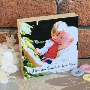 May include: A wooden photo block with a picture of a young child holding an older person's hand. The photo is surrounded by a white border and the text "I love you Grandad, Love Isla x" is printed below the photo.