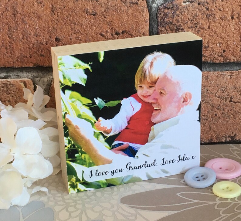 May include: A wooden photo block with a picture of a young child holding an older person's hand. The photo is surrounded by a white border and the text "I love you Grandad, Love Isla x" is printed below the photo.