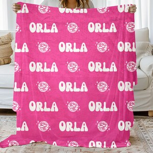 May include: A hot pink blanket with the name "ORLA" repeated in a white, retro font. The name is interspersed with white disco ball graphics. The blanket is held up in front of a white couch and a beige rug.