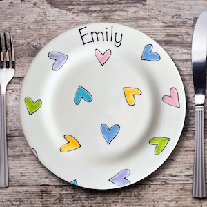 May include: A white ceramic plate with colorful heart designs and the name "Emily" in black script. A fork and knife are placed on either side of the plate on a wooden surface. The hearts are outlined in black and filled with various colors.