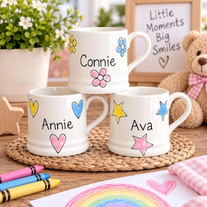 Op de afbeelding: Drie witte keramische mokken met oren, elk gepersonaliseerd met een naam in zwarte letters. De bovenste mok toont de naam Connie met kleurrijke bloemen. De onderste mokken tonen de namen Annie met hartjes en Ava met sterren als decoratie.
