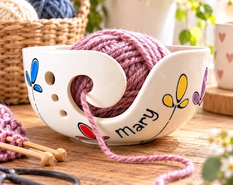 May include: A white ceramic yarn bowl featuring a spiral cutout, three small circular holes, and hand-painted colorful floral motifs. The name Mary is written in black script on the front, containing a ball of dusty rose yarn.