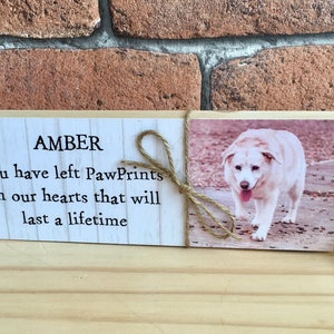 May include: A wooden memorial plaque with a white background and black text that reads "AMBER You have left PawPrints on our hearts that will last a lifetime". A photo of a white dog is on the right side of the plaque.