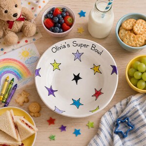 May include: A white ceramic bowl featuring the text Olivia's Super Bowl and a pattern of colourful hand-drawn stars in purple, yellow, red, blue, and black. The bowl is surrounded by a teddy bear, a bowl of berries, milk, crackers, and grapes.