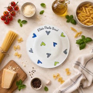 May include: A white ceramic pasta bowl featuring the text "Ella's Pasta Bowl" in black lettering, decorated with a pattern of small hand-drawn hearts in shades of blue, green, and grey scattered across the rim and the centre of the bowl.