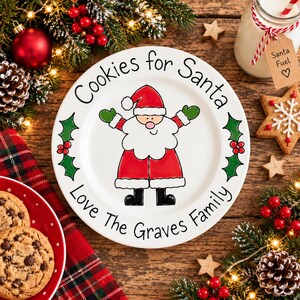 May include: A white ceramic plate featuring a cartoon Santa Claus in the center, surrounded by green holly leaves and red berries. The text reads "Cookies for Santa" at the top and "Love The Graves Family" at the bottom in black lettering.