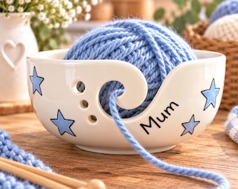 May include: A white ceramic yarn bowl featuring a spiral cutout for yarn, decorated with light blue stars and the word Mum in black script. A ball of light blue yarn sits inside the bowl, with the strand feeding through the spiral opening.