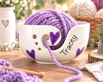 May include: A white ceramic yarn bowl featuring a spiral cutout, purple heart illustrations, and the name Tracey printed in black script. A large ball of purple yarn sits inside the bowl, with a strand feeding through the spiral opening.