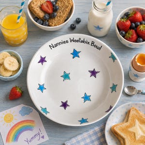 May include: A white ceramic bowl featuring the text Nannies Weetabix Bowl and a pattern of blue and purple stars. The bowl is surrounded by a glass of orange juice, a bowl of cereal with strawberries, a milk bottle, a bowl of sliced bananas, and toast.