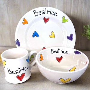 May include: A white ceramic dish set featuring a plate, bowl, and mug, each adorned with colorful heart designs and the name "Beatrice" in black script. The set is designed for dining.