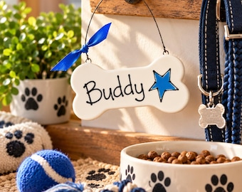 May include: A white bone-shaped ceramic sign with the name Buddy and a blue star printed on it, hanging from a black wire with a blue ribbon. The sign is displayed near a blue dog leash, a white bowl with black paw prints, and a blue tennis ball.