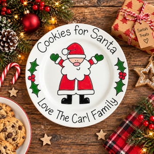 May include: A white ceramic plate featuring a cartoon Santa Claus in the center, with the text "Cookies for Santa" at the top and "Love The Carl Family" at the bottom, accented by green holly leaves and red berries on the left and right sides.
