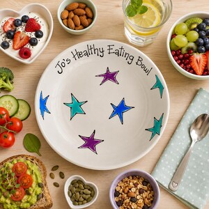 May include: A white ceramic bowl featuring the black text Jo's Healthy Eating Bowl and hand-painted purple, teal, and blue stars. The bowl is surrounded by various healthy food items including yoghurt with berries, almonds, cucumber, tomatoes, and granola.