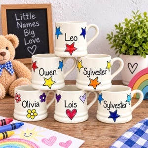 Op de afbeelding: Zes witte keramische mokken met oren, elk voorzien van een unieke naam in zwarte letters en handgetekende motieven zoals sterren, hartjes en bloemen. De namen op de mokken zijn Leo, Owen, Sylvester, Olivia, Lois en Sylvester.