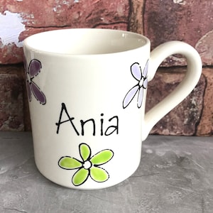 May include: White ceramic mug with a black handle. The mug features a green, purple, and white flower design and the name "Anya" written in black.