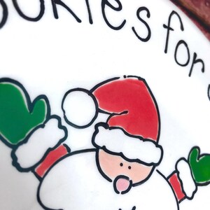 May include: A white ceramic plate featuring a hand-drawn illustration of Santa Claus wearing a red hat and green mittens. The text "okies for" is printed in black ink along the top edge of the plate. The design uses bold black outlines.