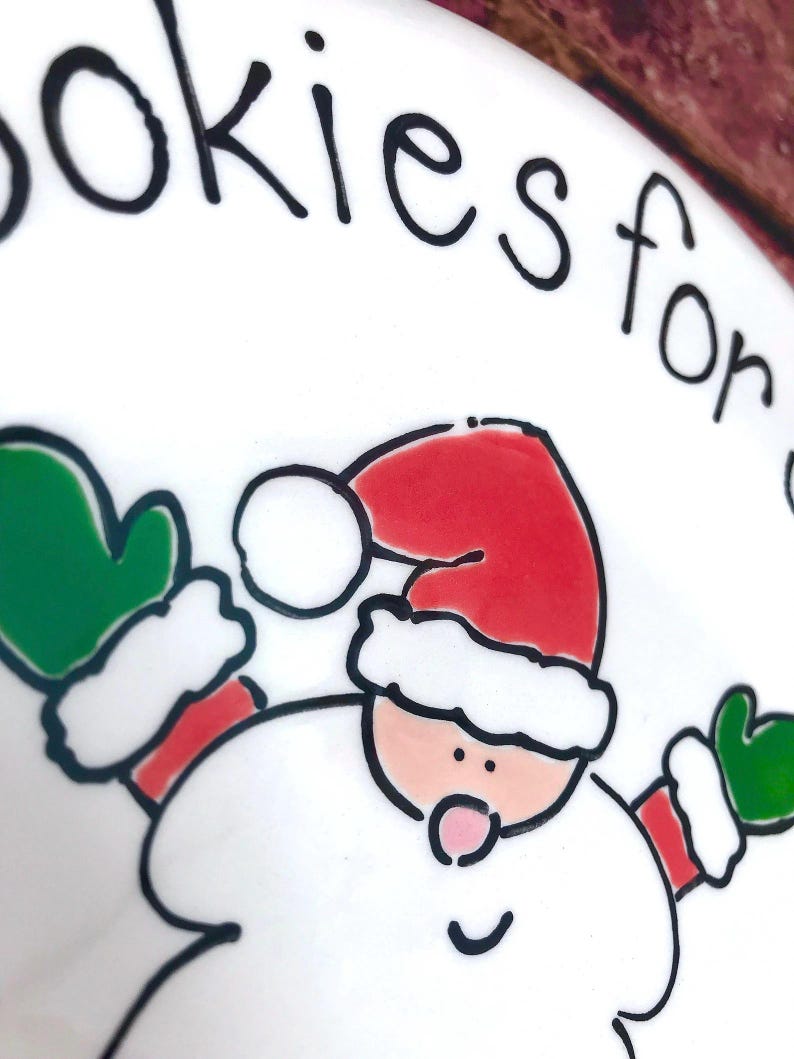 May include: A white ceramic plate featuring a hand-drawn illustration of Santa Claus wearing a red hat and green mittens. The text "okies for" is printed in black ink along the top edge of the plate. The design uses bold black outlines.