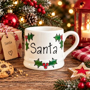 May include: A white ceramic mug featuring the word Santa written in black script, decorated with green holly leaves and red berries. The mug has a rounded handle and a smooth finish, with the design printed on the front surface of the vessel.