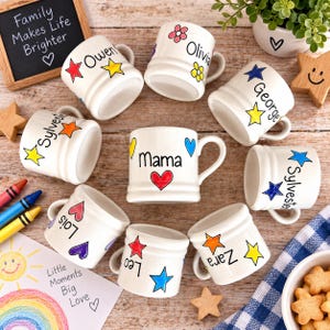 Op de afbeelding: Een verzameling witte keramische mokken met handgeschilderde kleurrijke sterren, harten en bloemen. Elke mok is gepersonaliseerd met een naam in zwarte letters, waaronder Owen, Olivia, George, Sylvester, Lois, Mama, Leo en Zara.