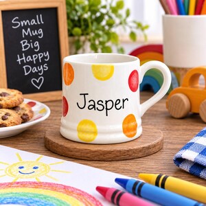 Op de afbeelding: Een witte keramische mok met de naam Jasper in zwarte letters, versierd met kleurrijke gele, oranje en rode stippen. De mok heeft een gebogen handvat en staat op een ronde houten onderzetter, een klassiek ontwerp voor dagelijks gebruik.