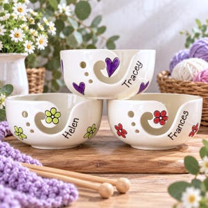 May include: Three white ceramic yarn bowls with a spiral cutout for yarn. The top bowl features purple hearts and the name Tracey. The bottom left bowl has light green flowers and the name Helen. The bottom right bowl has red flowers and the name Frances.