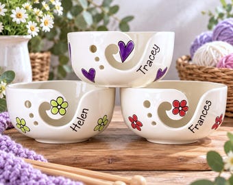 May include: Three white ceramic yarn bowls with a spiral cutout for yarn. The top bowl features purple hearts and the name Tracey. The bottom left bowl has light green flowers and the name Helen. The bottom right bowl has red flowers and the name Frances.