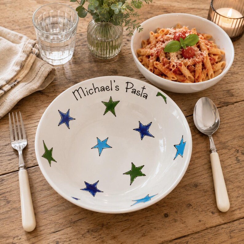 May include: A white ceramic bowl featuring the text Michael's Pasta and a pattern of blue, light blue, and green stars. The bowl is accompanied by a silver fork and a silver spoon, both with cream-coloured handles, arranged on a wooden surface.