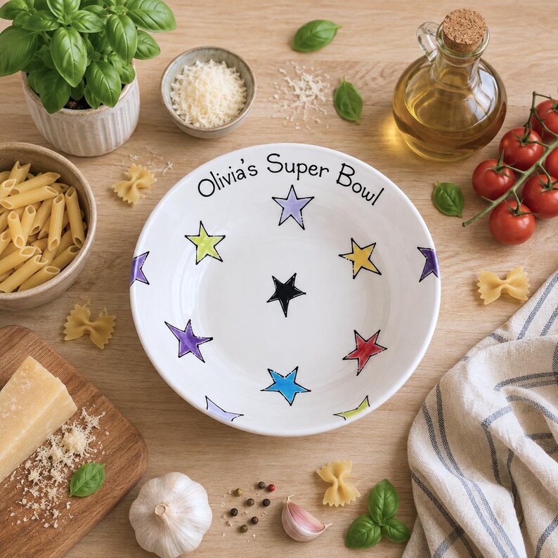 May include: A white ceramic bowl featuring the black text "Olivia's Super Bowl" and a pattern of multi-coloured stars in shades of purple, yellow, red, blue, and green scattered across the interior surface of the bowl.