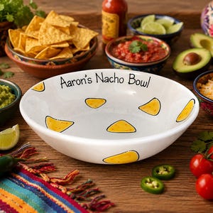 May include: A white ceramic bowl with a hand-painted yellow nacho chip pattern and the black text Aaron's Nacho Bowl printed along the inner rim. The bowl has a wide, shallow design with a smooth, glossy finish and a rounded edge.