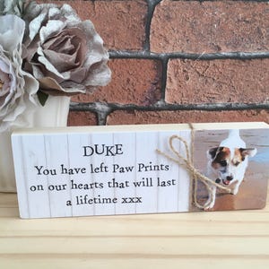 May include: A wooden memorial plaque with a white background and black text that reads "DUKE You have left Paw Prints on our hearts that will last a lifetime xxx". A photo of a dog is on the right side of the plaque.