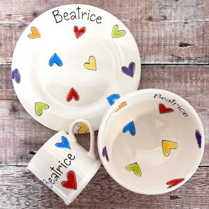 May include: A personalized children's dinnerware set with a white background and colorful hearts. The set includes a plate, bowl, and mug, all featuring the name "Beatrice" and colorful hearts.