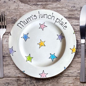 May include: White ceramic plate with the text "Mum's lunch plate" and colorful star designs. A fork and knife are placed on a wooden surface, flanking the plate. The stars are in shades of pink, blue, yellow, and green.